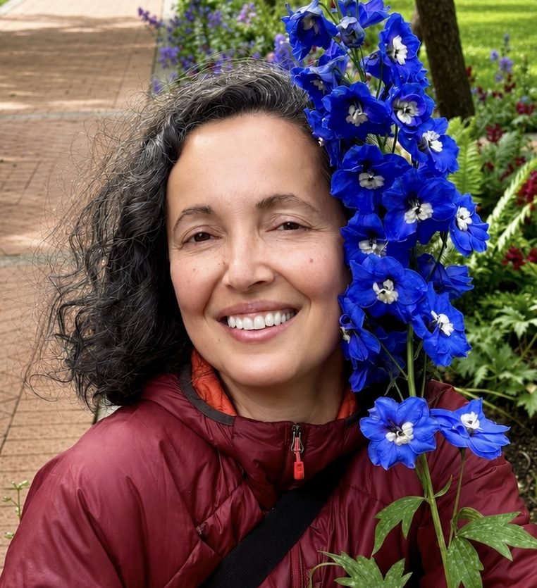 Marion smiling outdoors beside a tall stem of blue delphinium flowers